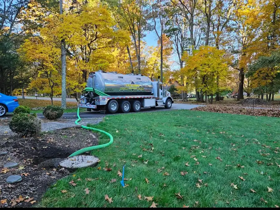 Black Diamond tanker truck parked on a lawn with a green suction hose running to an open septic tank, autumn trees behind